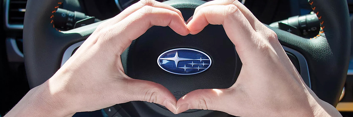 Person holding a heart with their hands over the subaru logo inside a car